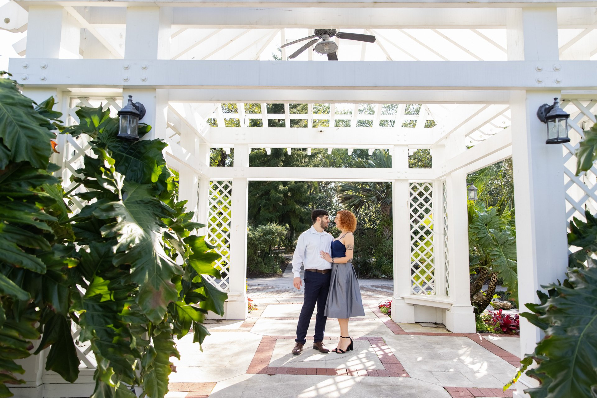 A couple standing under a white pergola with lattice details, surrounded by green foliage in an outdoor garden setting. They are looking at each other, creating a romantic scene.