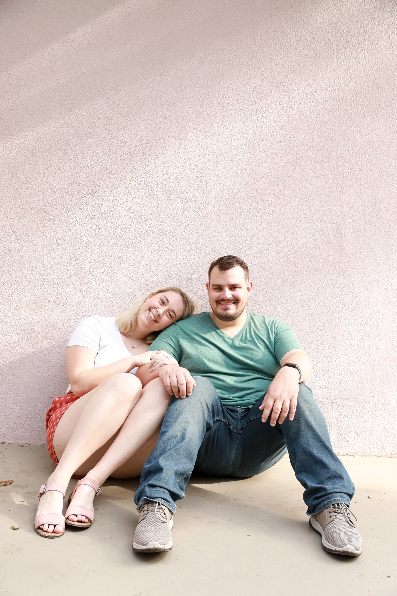 A couple sitting on a floor against a textured wall, the woman leaning her head on the man's shoulder, both smiling.