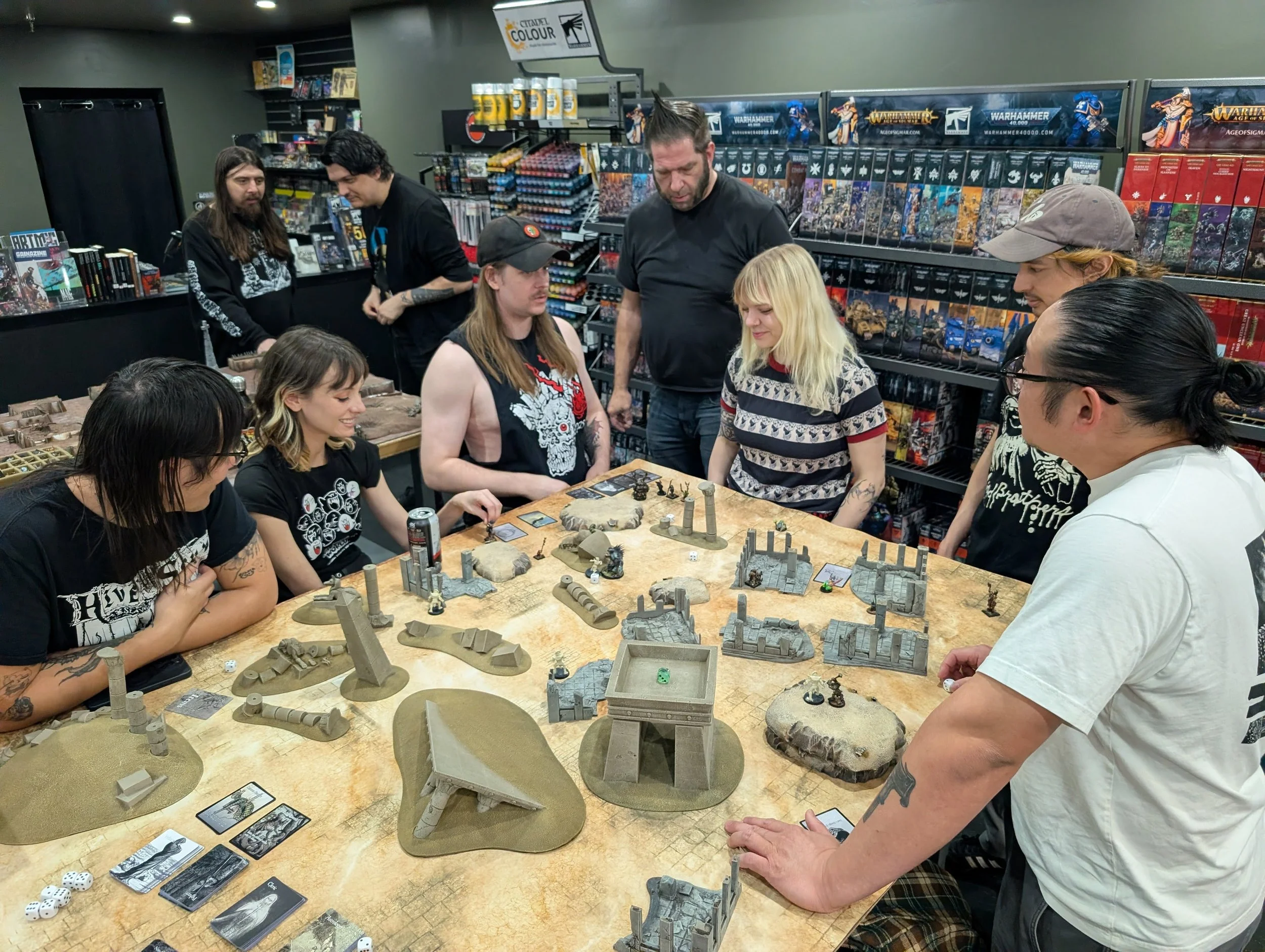A group of people playing a tabletop miniature wargame at a table in a game store. There are miniatures, terrain pieces, cards, and dice on the table. Shelves behind them display boxed games and figurines.