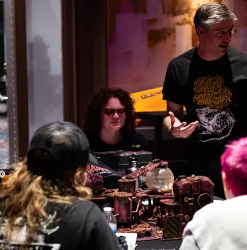 A group of people sitting around a tabletop lab with miniature terrain and a crystal ball, with one person in glasses and headphones, another with pink hair, and a man standing and talking.