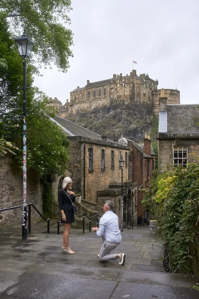 A man kneeling on one knee proposing to a woman on a rainy city street with historic buildings and a castle on a hill in the background.