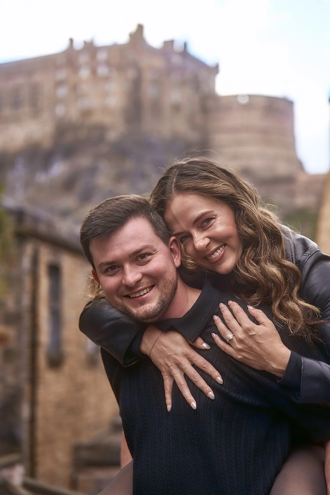 A young woman and a young man sitting together on a balcony, with the woman leaning her head on the man's shoulder, smiles at the camera, with a cityscape of historic rooftops in the background.