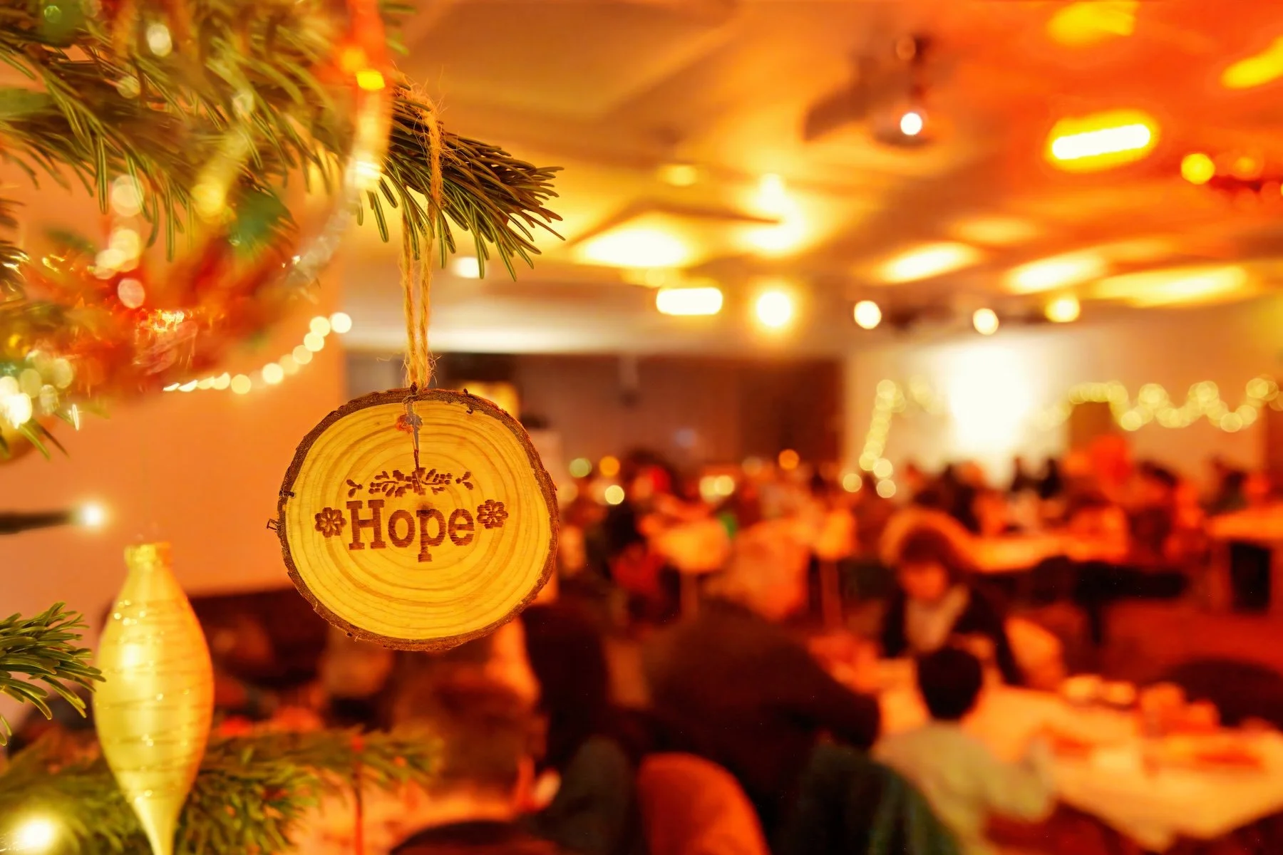 A wooden ornament with the word 'Hope' engraved, hanging from a Christmas tree branch in a warmly lit indoor setting filled with many people.