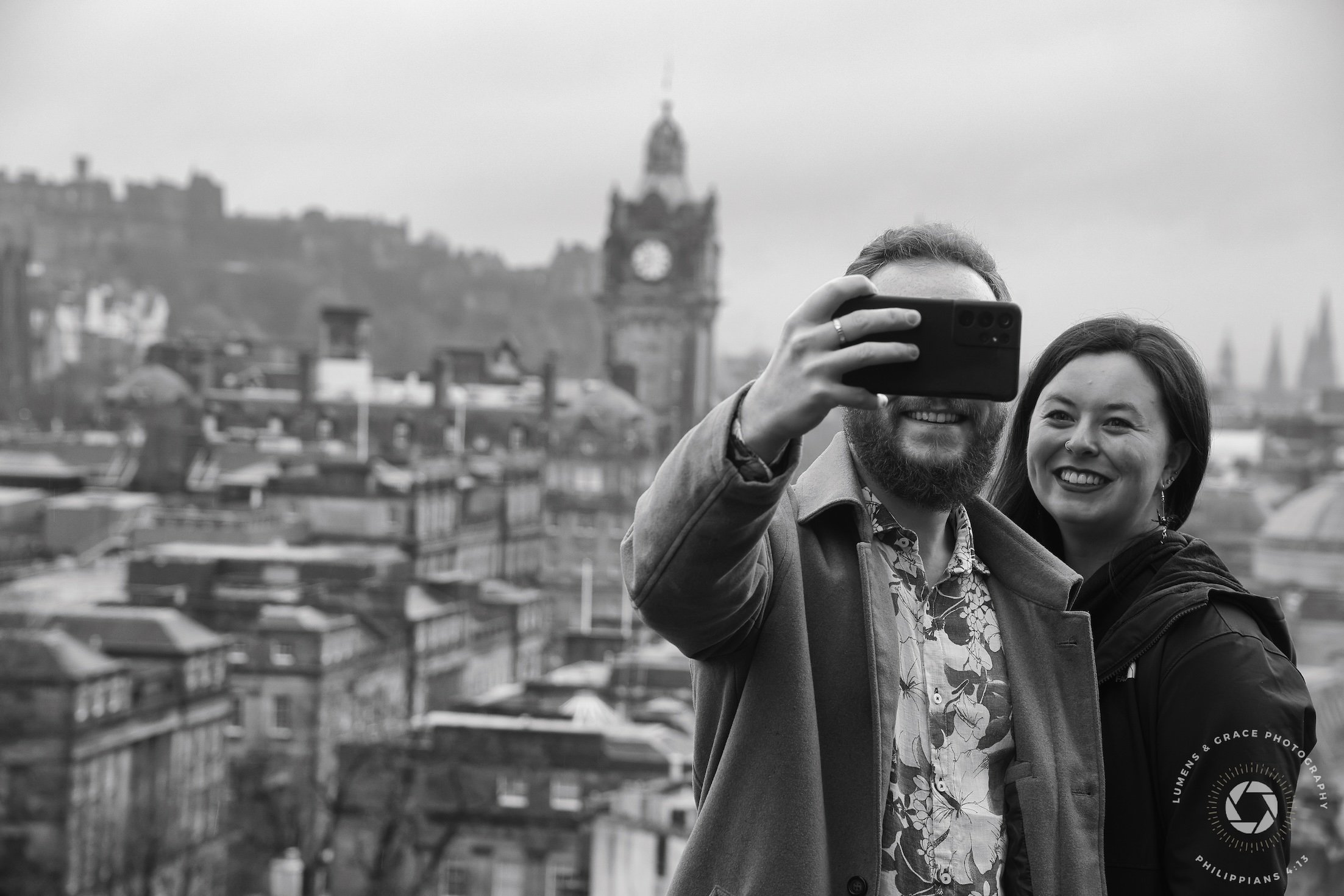 A man and woman taking a selfie with a scenic cityscape in the background. The image is in black and white, showing historical buildings and a clock tower in the distance. The couple is smiling and the man is holding a smartphone.