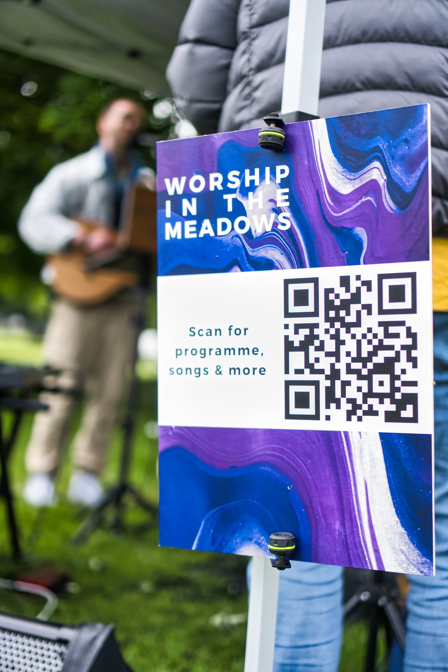 Sign with a QR code for 'Worship in the Meadows' event, blurred background of a person playing guitar.