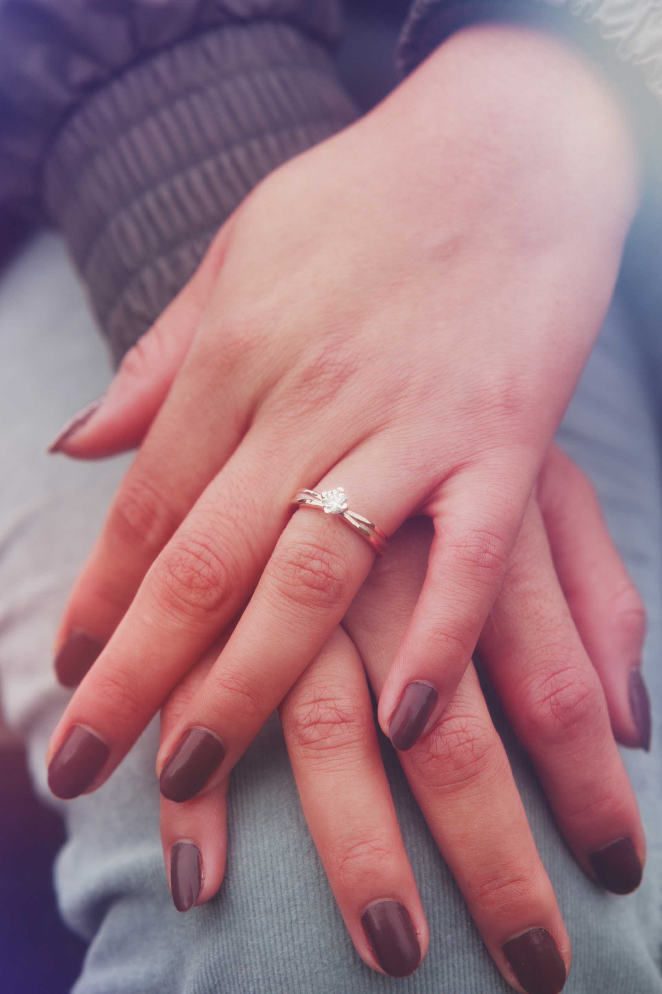 Close-up of two hands interlocked, one showing a diamond engagement ring, both with painted nails, against a background of a grey fabric.