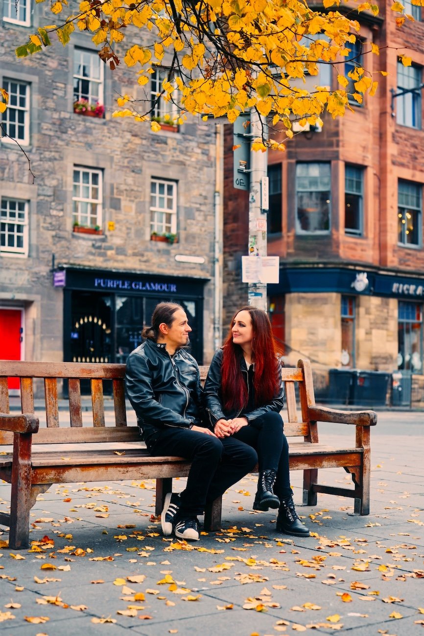 A man and a woman sitting on a wooden park bench, holding hands, surrounded by fallen autumn leaves, with buildings and trees displaying yellow foliage in the background.