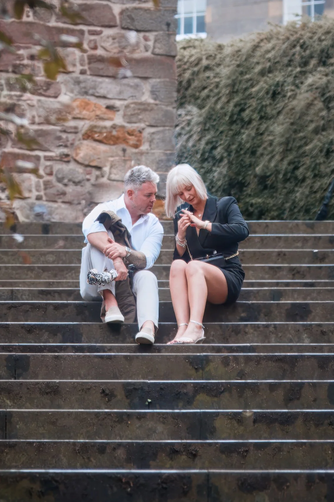 A man and a woman sitting on outdoor steps, looking at a phone together, with a child standing next to the man, behind the woman, and a stone wall and bushes in the background.