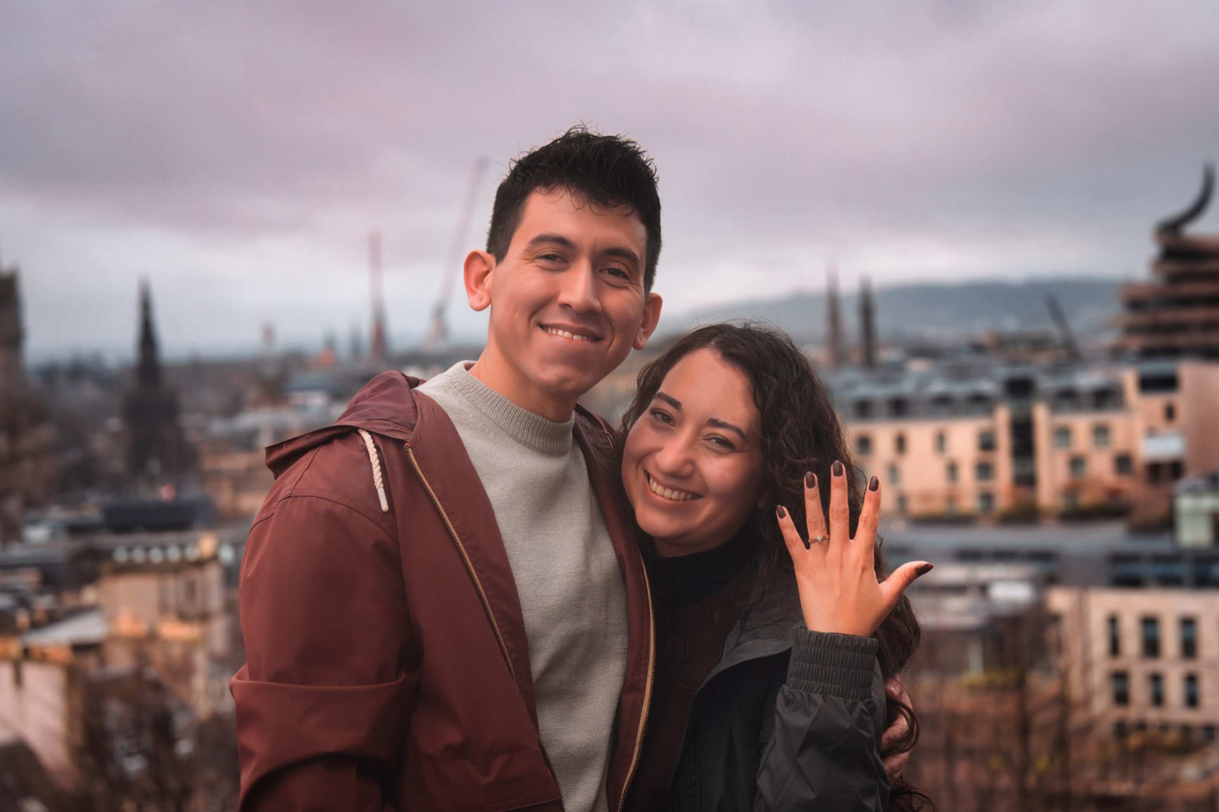 Two smiling young people, a man and a woman, standing close together outdoors during cloudy weather. The man has short dark hair and is wearing a maroon jacket over a gray sweater. The woman has curly dark hair, tattoos on her left hand, and is weari