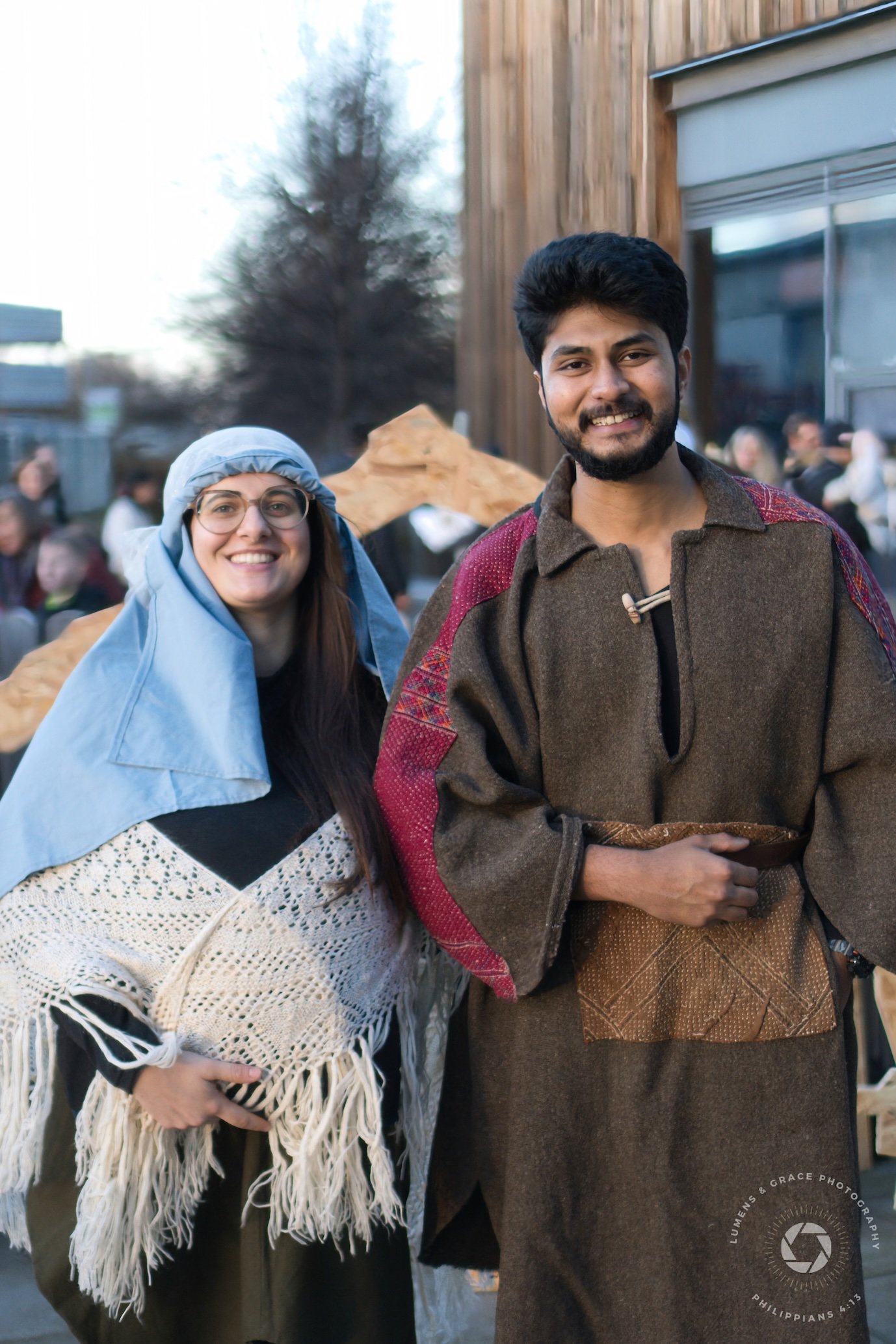 Two people dressed in Biblical costumes, smiling outdoors, with a wooden prop in the background.
