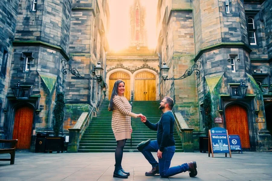 A couple in front of a historic stone building with stairs, one person kneeling and proposing to the other, holding hands and smiling.
