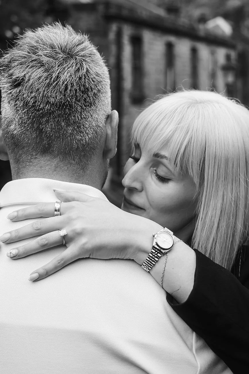 A woman with platinum blonde hair embracing a man from behind, with her eyes closed, wearing rings and a watch, in front of a blurred building background.