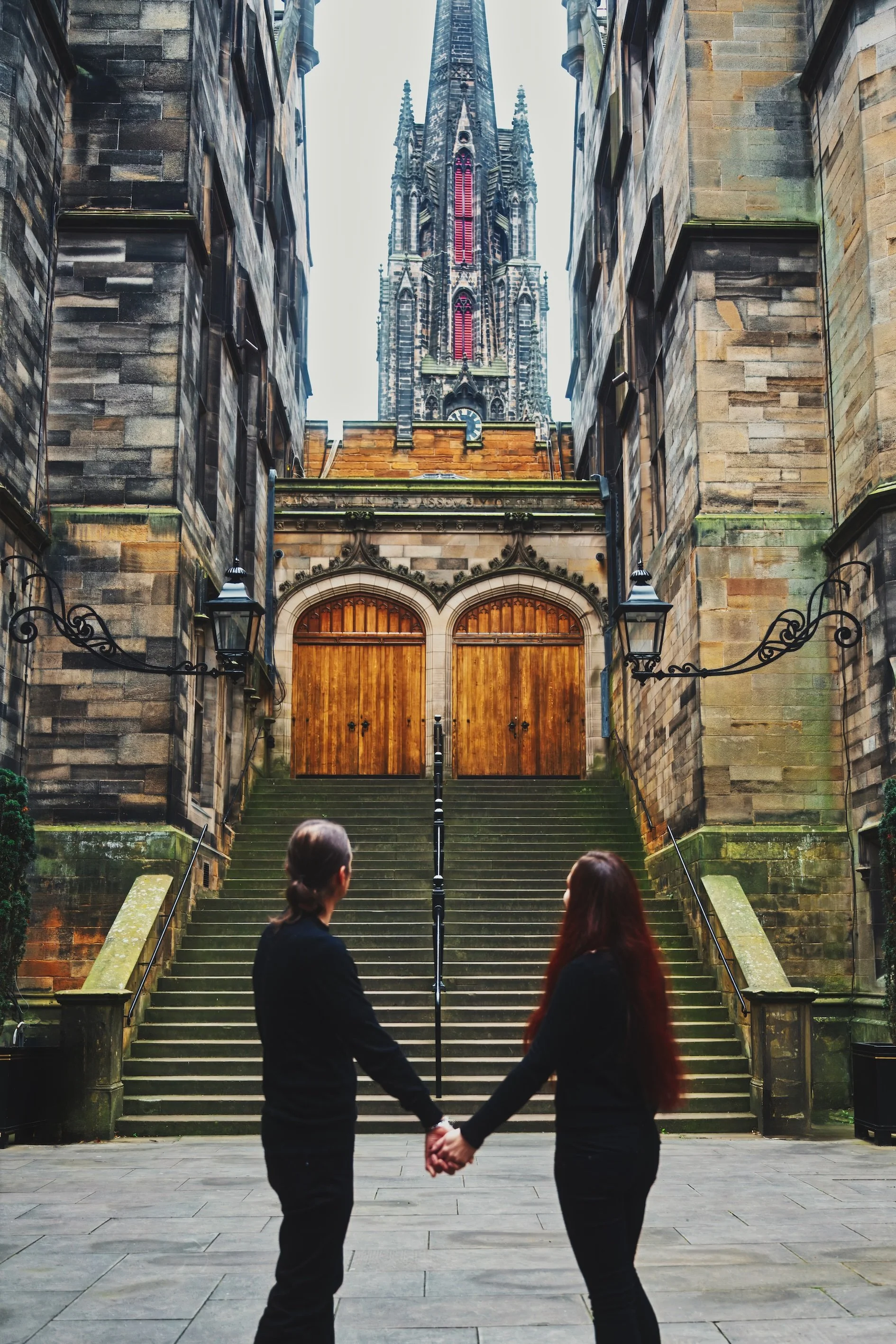 Two women holding hands in front of a staircase leading up to a gothic cathedral with tall spires and intricate detailed architecture.