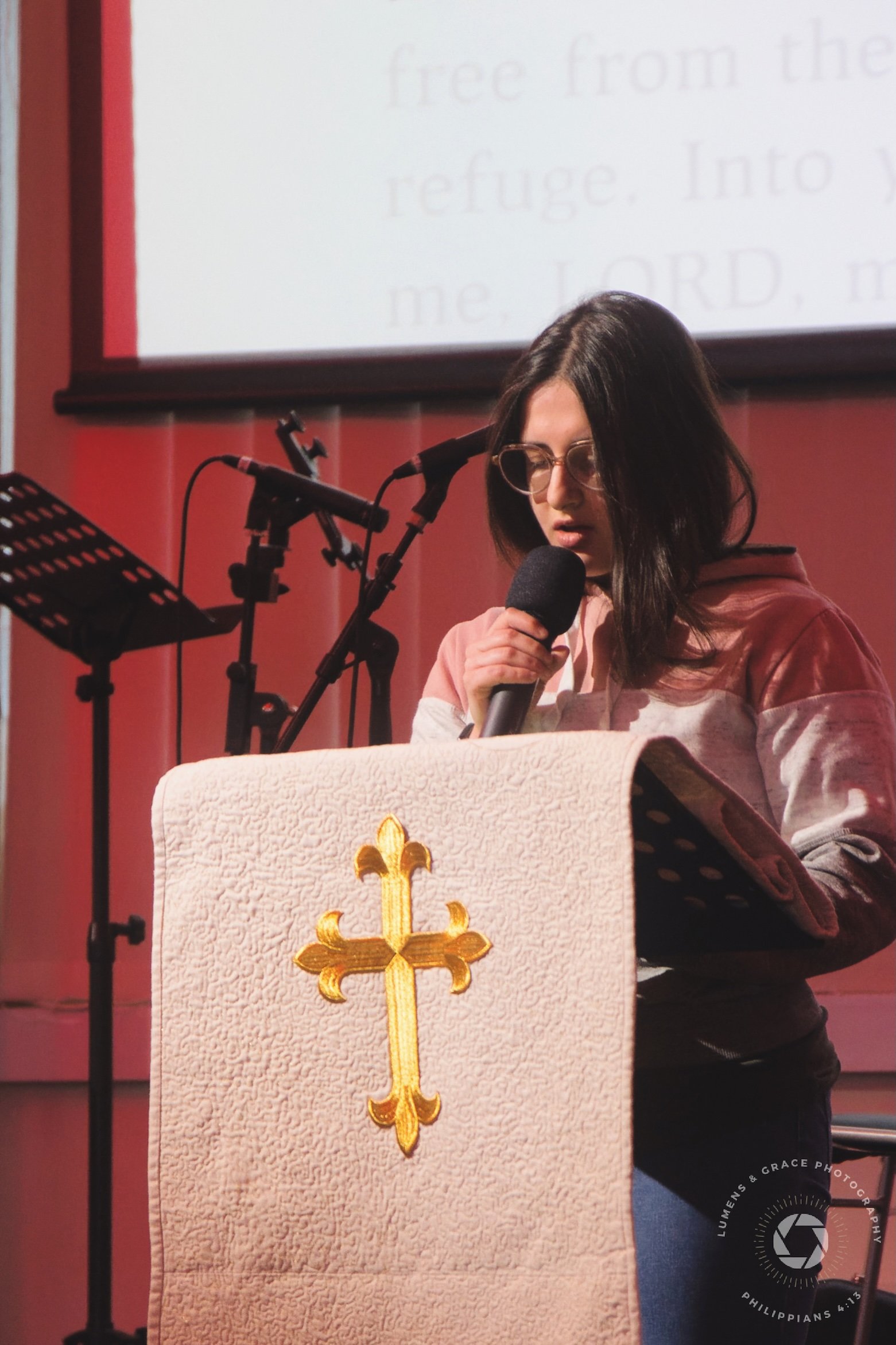 Person standing at a lectern with a microphone, reading from a pulpit adorned with a golden cross. Text is projected on a screen in the background.