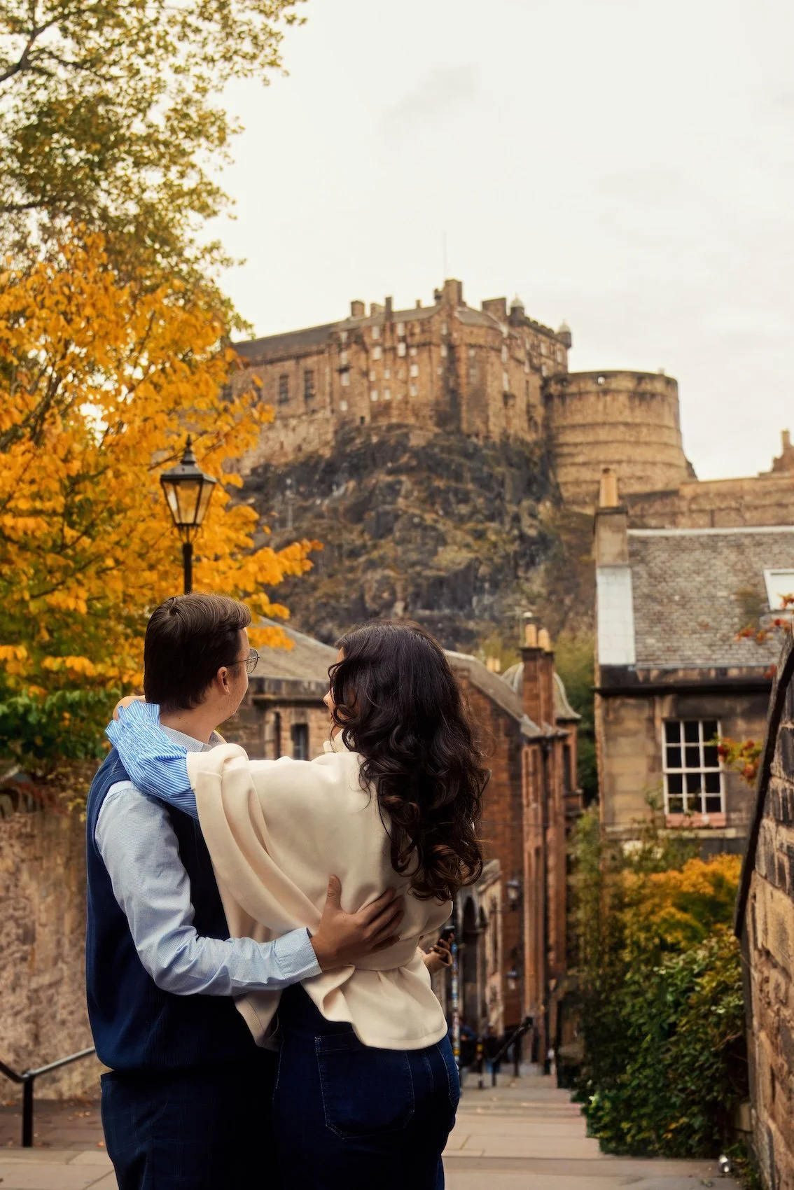 A couple hugging and looking at each other on a street with a castle on a hill in the background, autumn trees with yellow leaves, and old buildings.