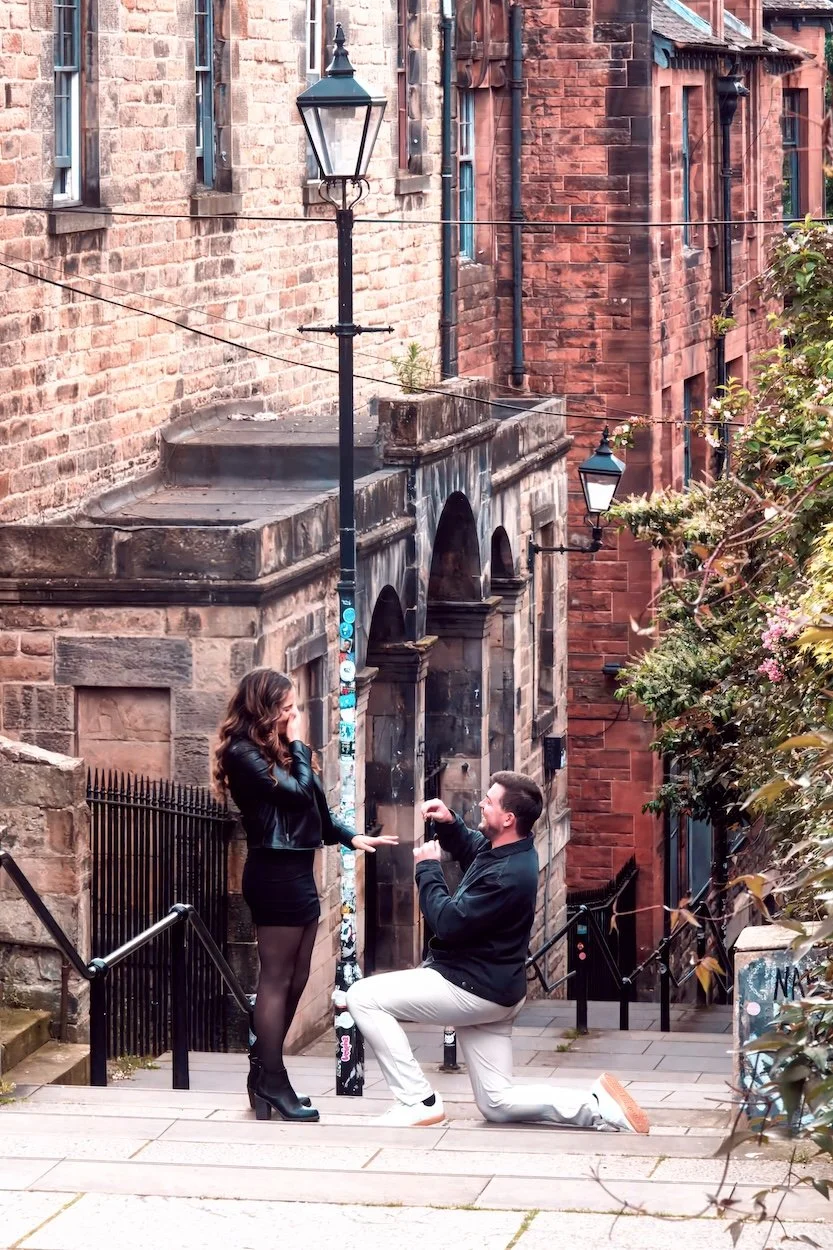 A man proposing marriage to a woman on a city street, with buildings made of red bricks and a lamppost in the background.