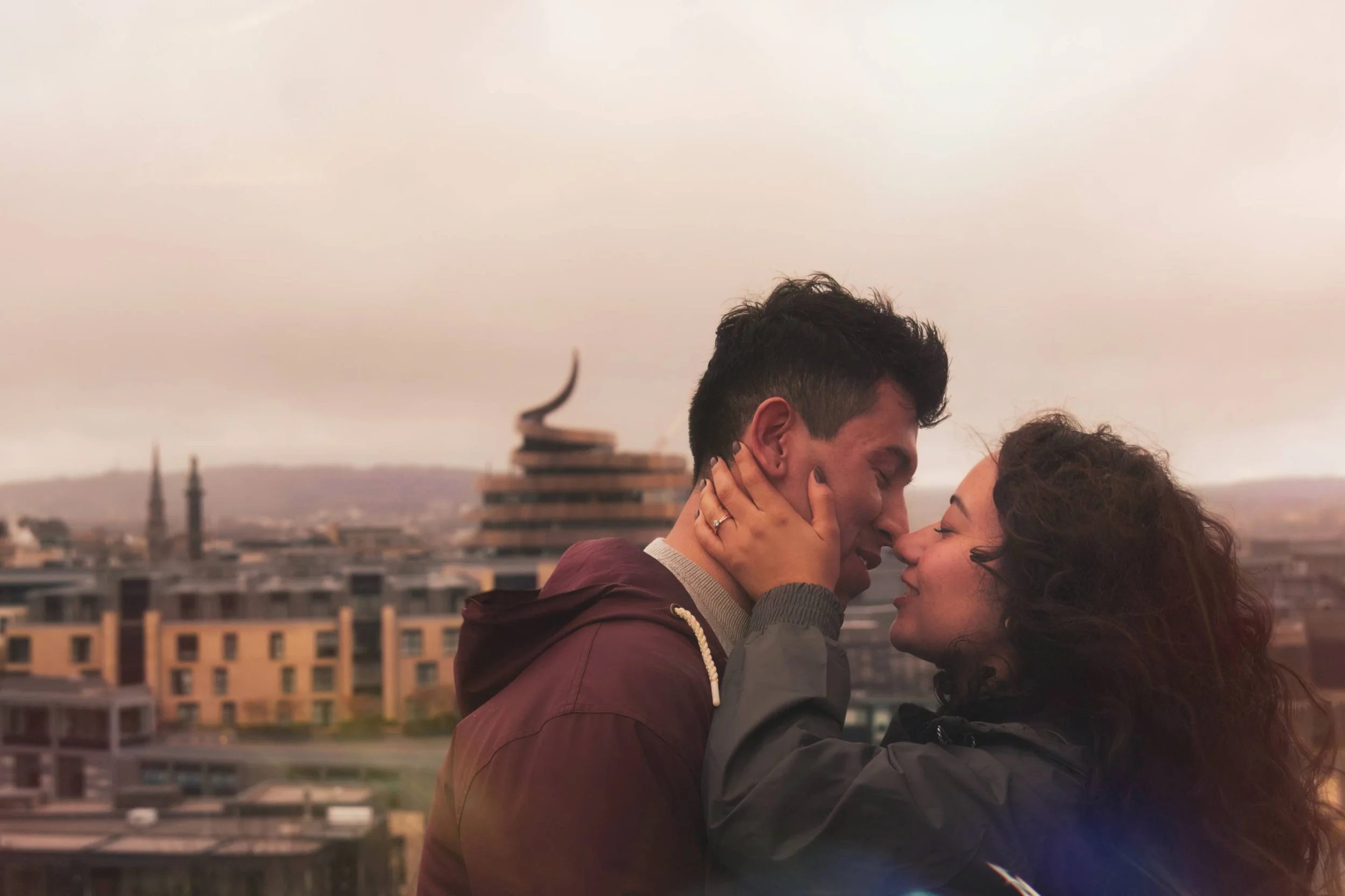 A couple stands close together, touching foreheads and smiling on a rooftop with city buildings and a cloudy sky in the background.