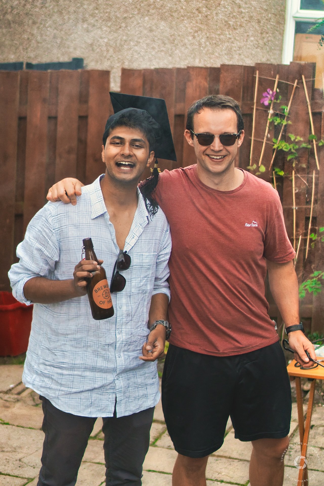 Two men smiling at an outdoor gathering, one wearing a graduation cap and holding a beer bottle.