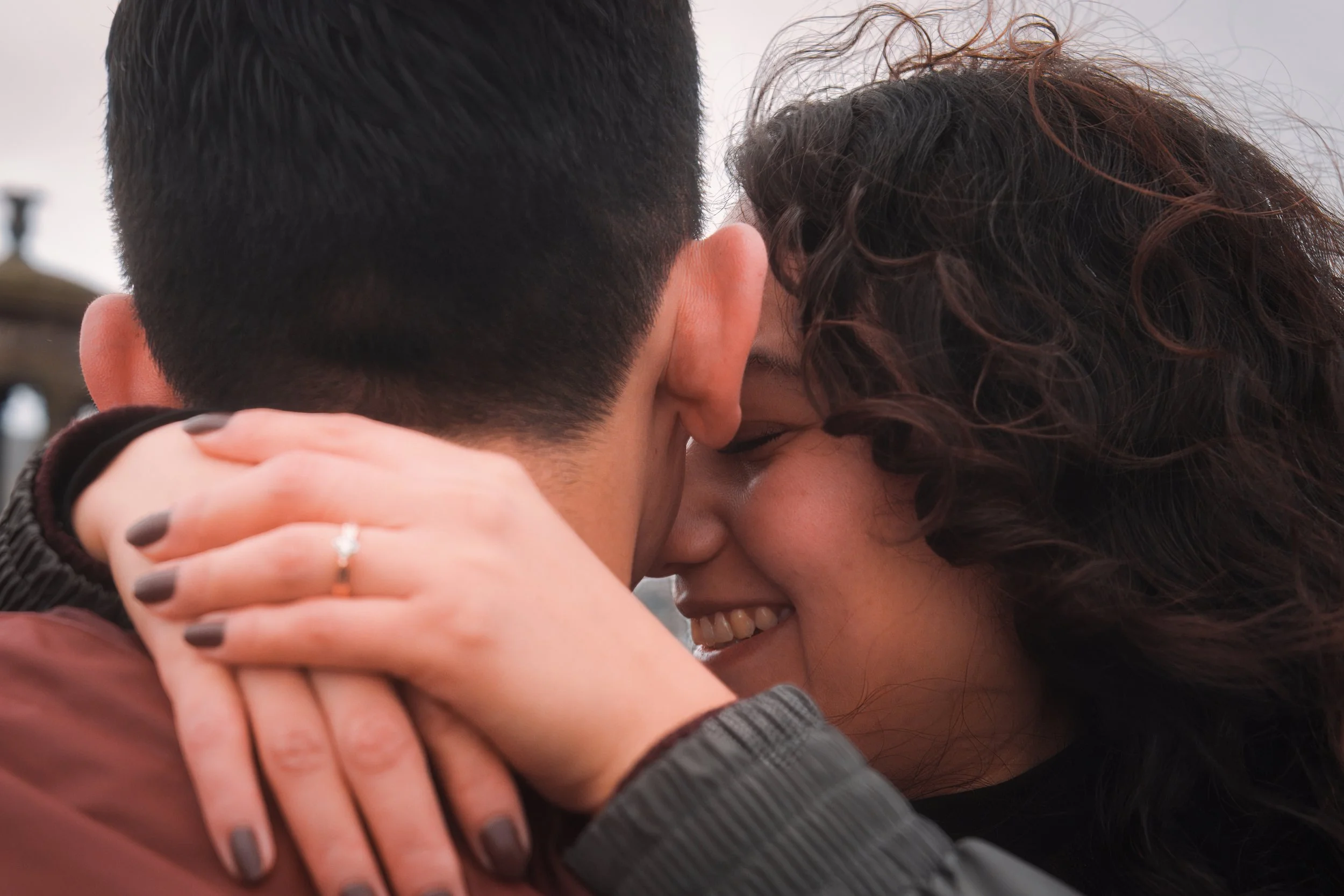 A close-up of two people sharing a kiss, with their faces pressed close and their eyes closed, smiling. One has curly hair, and the other has short dark hair. Their hands are intertwined around each other's necks.