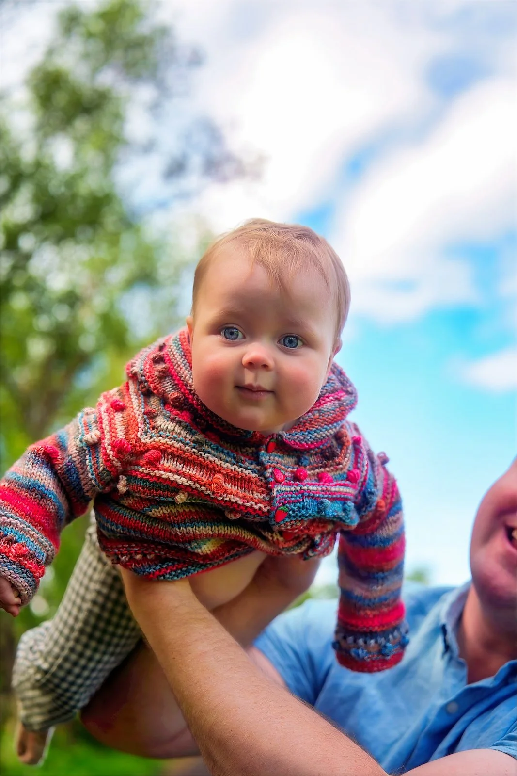 A baby with blue eyes and light brown hair wearing a colorful striped sweater, being held up outdoors by a person. There are green trees and a partly cloudy sky in the background.