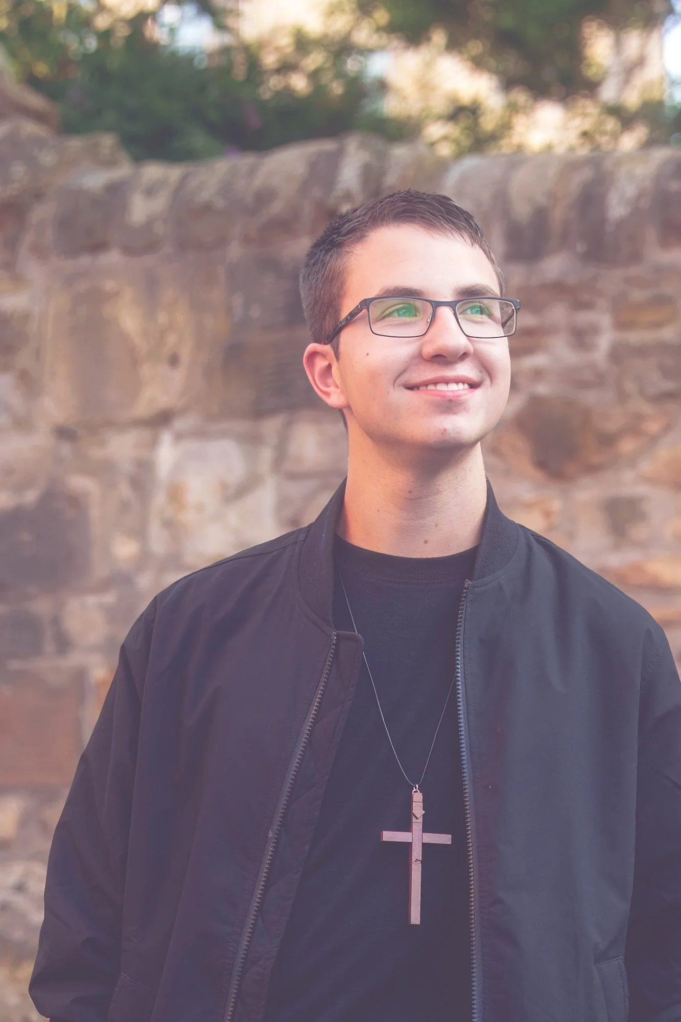 A young man with short dark hair, glasses, and a big cross necklace standing outdoors in front of a stone wall, smiling and looking to the side.