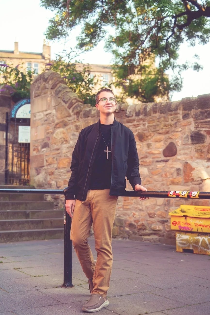 A young man with short dark hair, glasses, and a cross necklace is standing outdoors near a brick wall, holding onto a metal railing, smiling and looking up. He is wearing a black bomber jacket, black shirt, tan pants, and tan shoes. There is graffiti on a yellow box to his right, and trees and buildings in the background.
