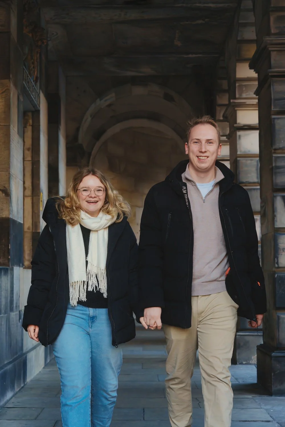 A smiling couple holding hands and walking outdoors in front of a stone building, dressed in casual winter clothing.