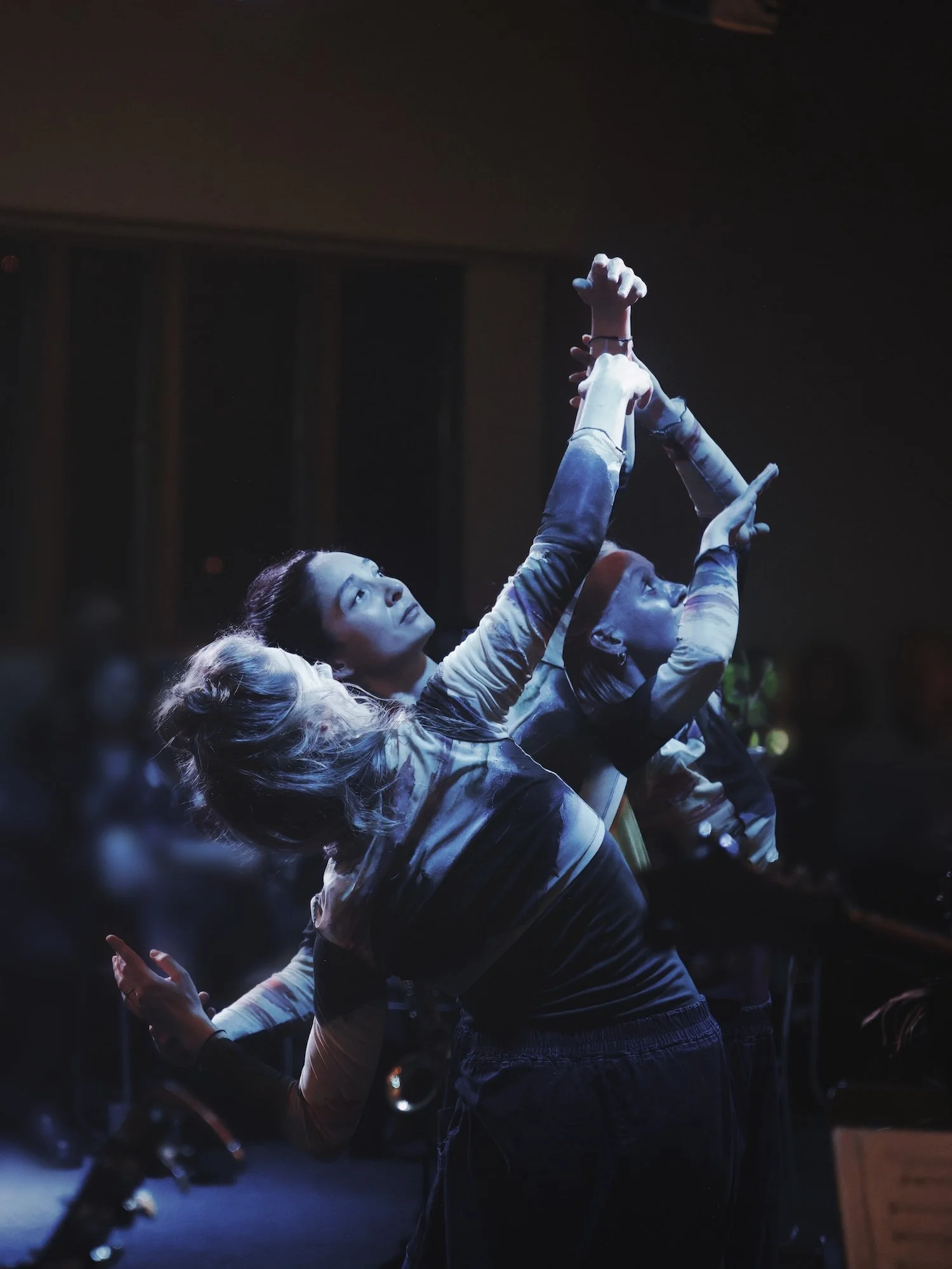 Group of dancers in a dark setting, performing with arms raised, illuminated by stage lighting.