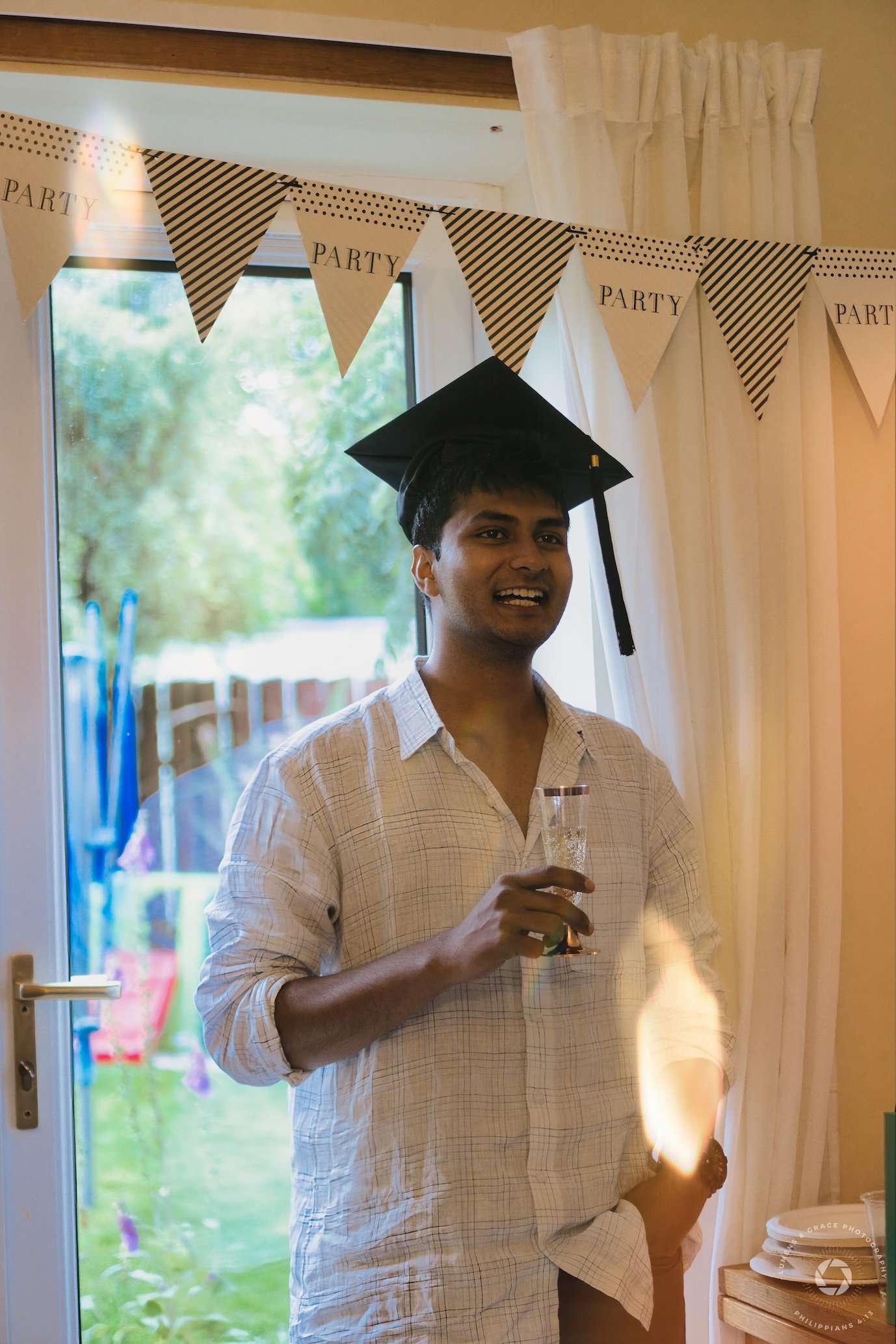 Person wearing a graduation cap, holding a glass, standing near a window with "Party" banners hanging above.