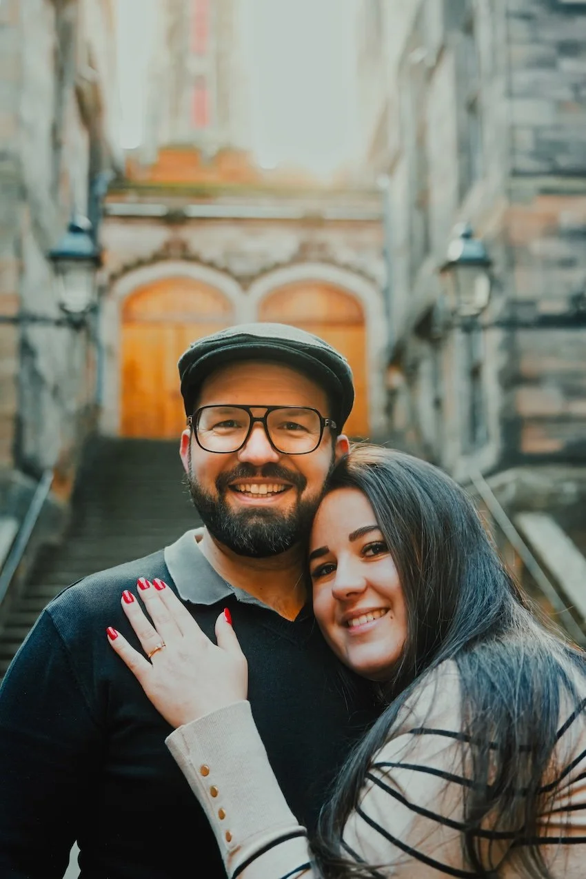 A smiling couple hugging each other on outdoor staircase with rustic brick and stone walls behind them.