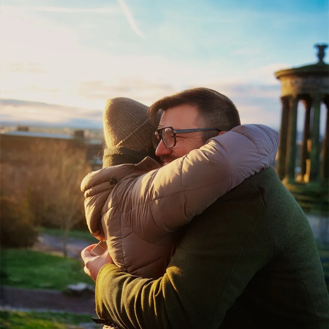 Two people hugging outdoors during sunset, with a scenic view and a historic structure in the background.