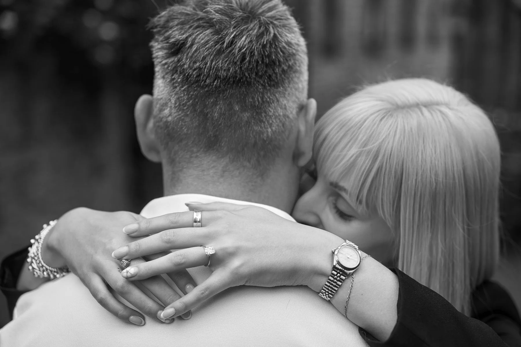 A black and white photograph of a couple embracing closely. The person on the left has short hair and is wearing a light-colored top, with their head turned away from the camera. The woman on the right with blond hair is embracing the other person, h