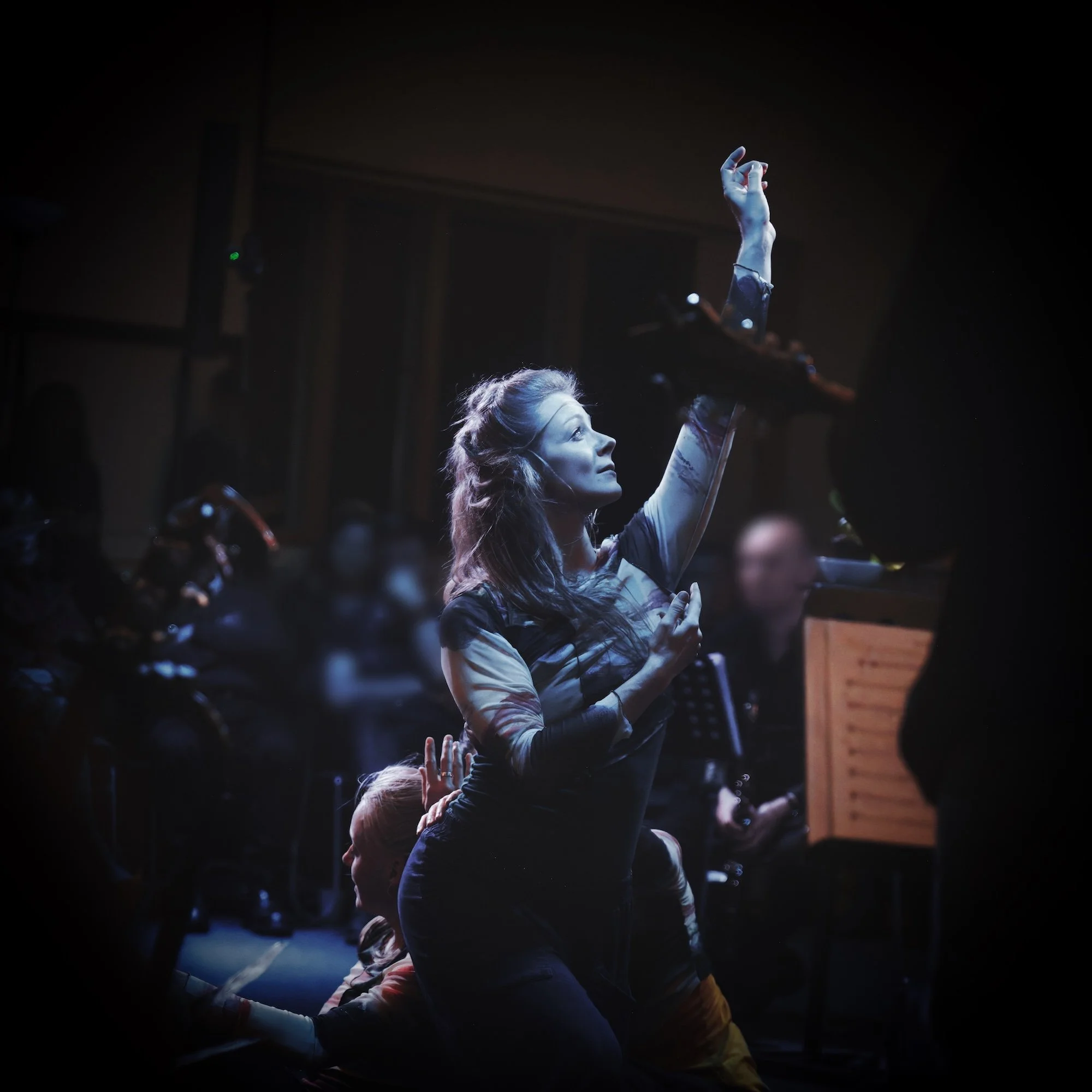 A woman participating in a performance or dance, with her arm raised and hand making a gesture, in a dark setting with surrounding people and stage equipment.
