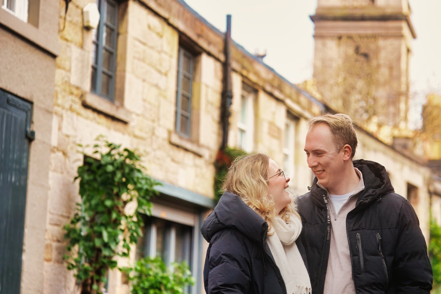 A happy couple smiling and looking at each other outdoors, with historic stone buildings in the background.