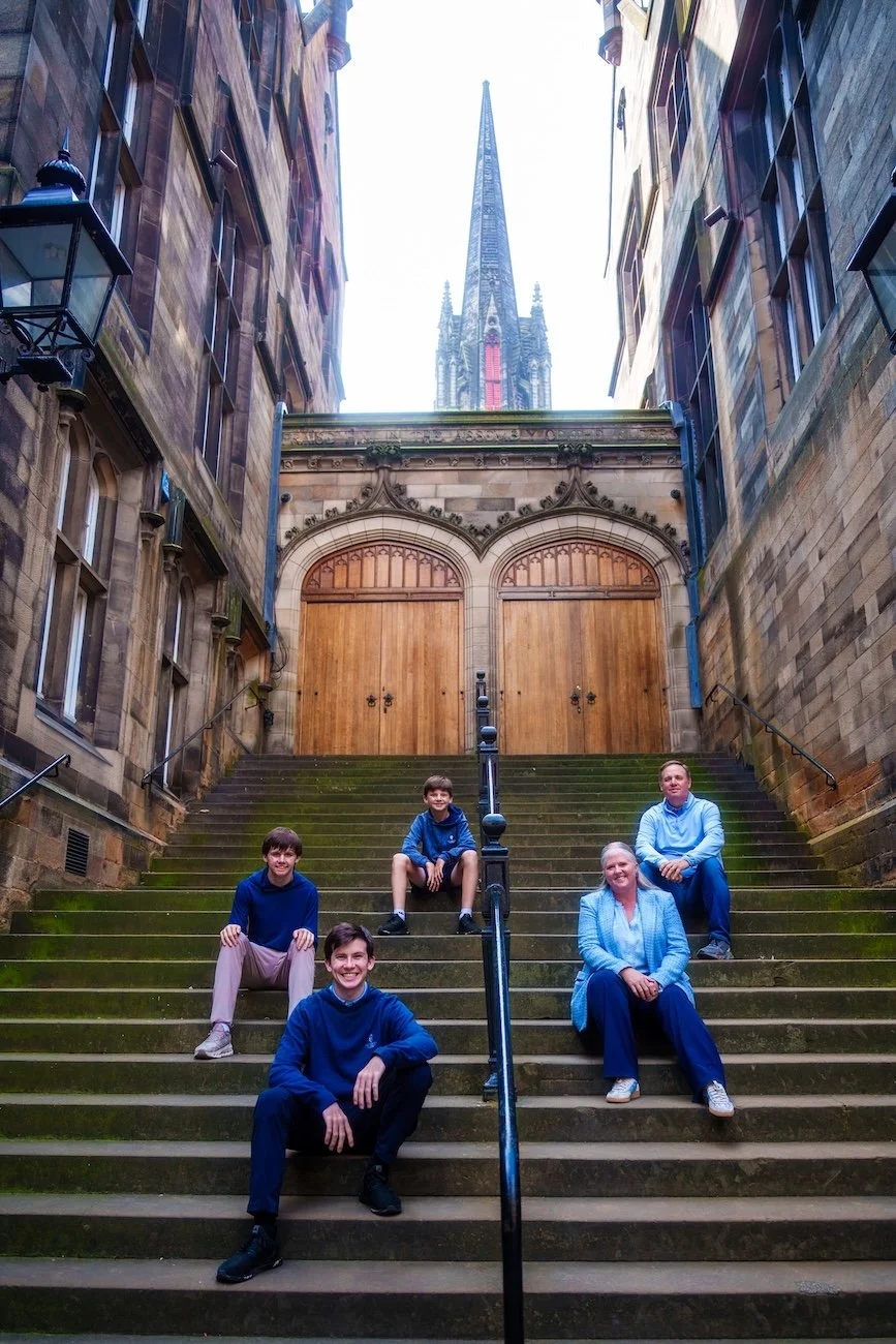 A group of five people, four young men and one woman, sitting on moss-covered stairs outside in front of a historic stone building with large wooden doors and an ornate archway, with a tall church steeple in the background.