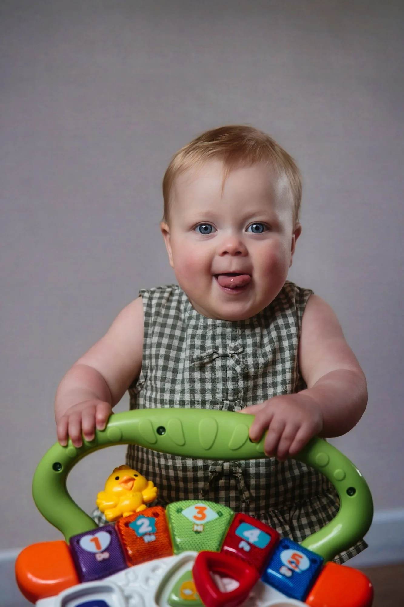 A young child with red hair and blue eyes sticking out their tongue, holding onto a green activity center with colorful blocks and toys, including a yellow rubber duck and numbered pieces.