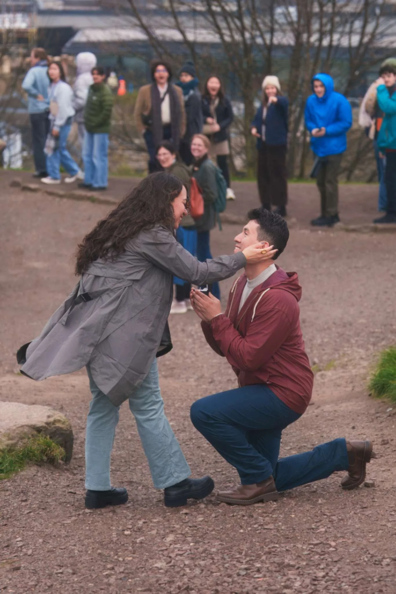 A man is kneeling on one knee holding a woman's face, while the woman is smiling and holding the woman's face with both hands. There are onlookers in the background, some smiling and observing the scene at Calton Hill.
