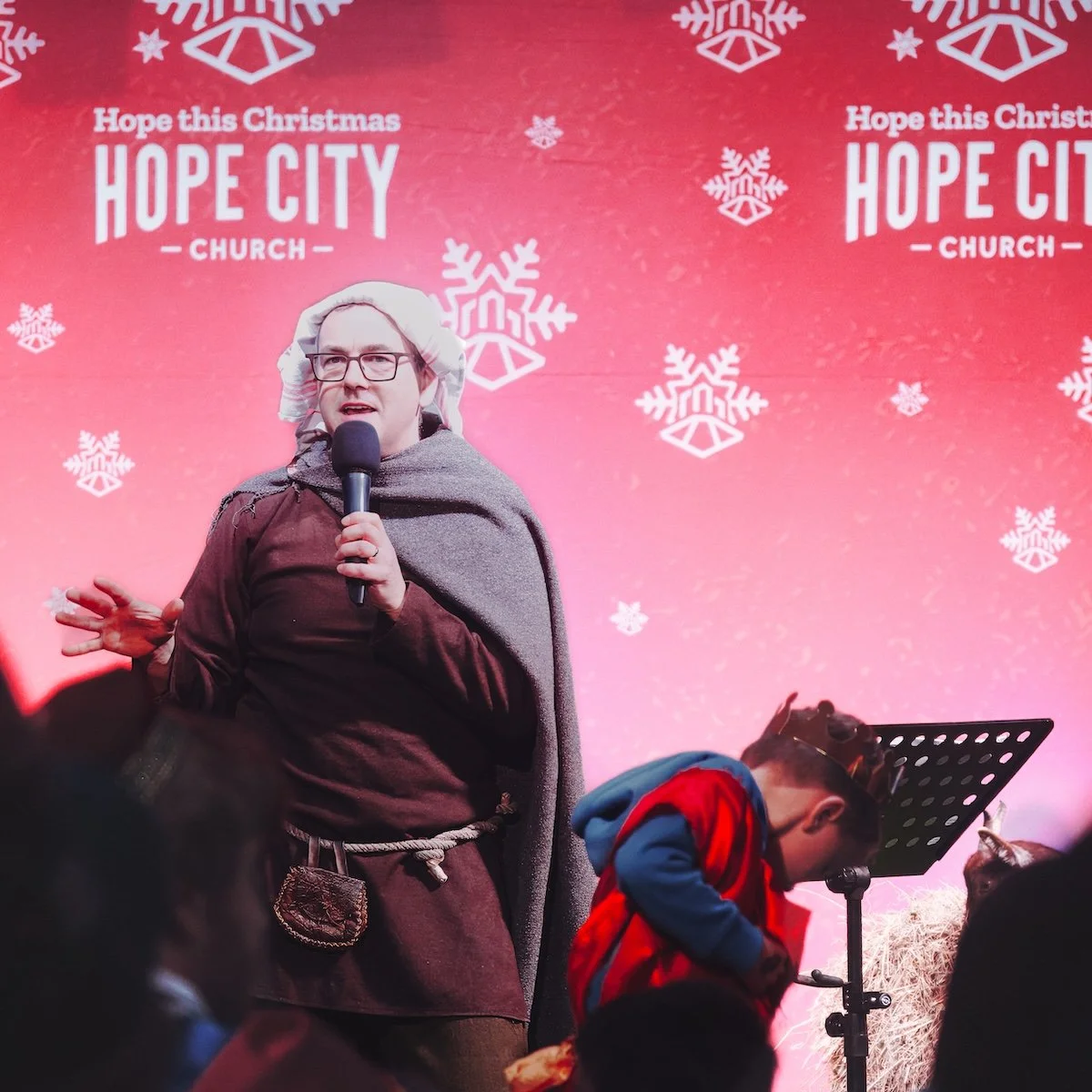 A person dressed as a historical or biblical figure holding a microphone and speaking in front of a festive Christmas backdrop with snowflakes. The backdrop features the words 'Hope this Christmas Hope City Church' with a pink and red gradient backgr