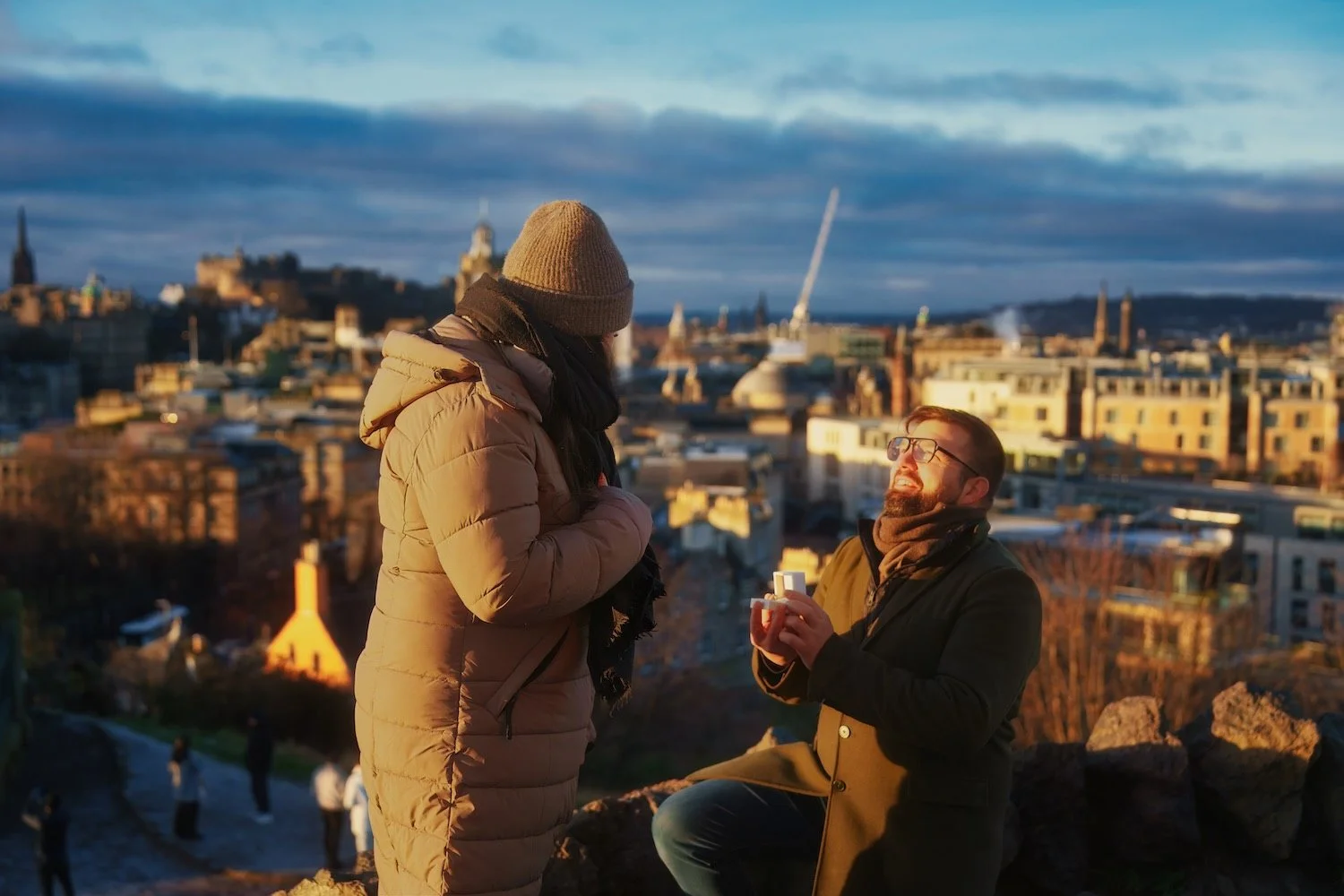 A man proposes marriage to a woman outdoors during sunset in a city with historic buildings, while the woman reacts happily.