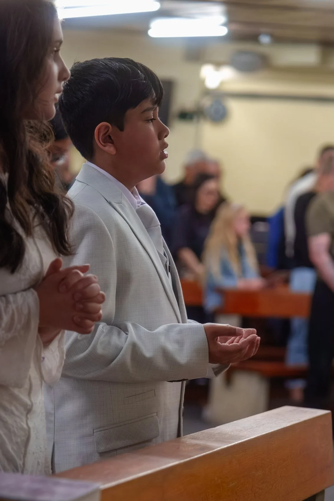 A young boy in a light-colored suit standing with hands clasped in prayer during a religious service, surrounded by other congregants in a church.