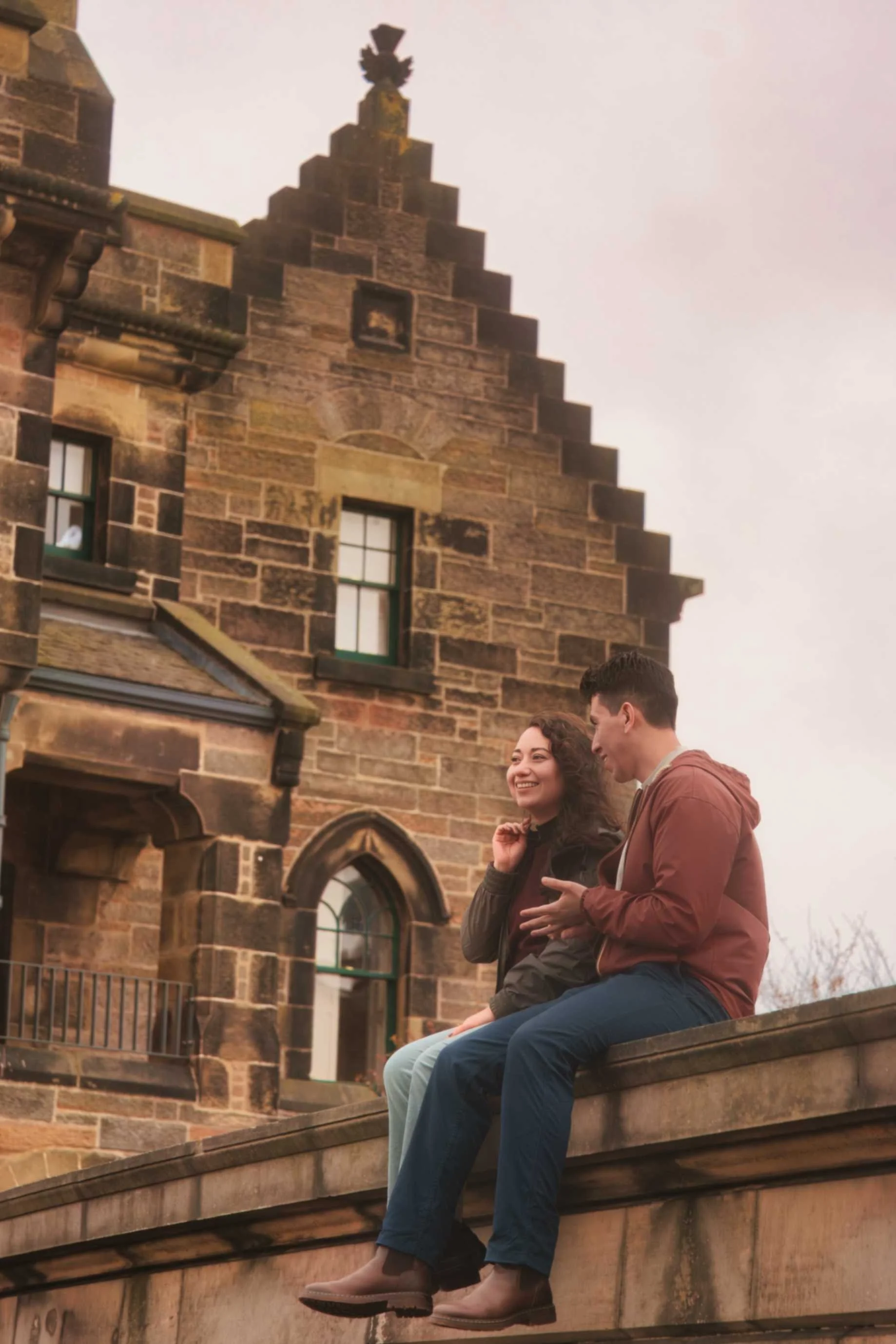 A man and woman sit on a wooden ledge in front of a historic stone building, smiling and talking. The building features gothic architecture with pointed arches and tall windows, and is made of reddish-brown stone. Some windows have small railings, an