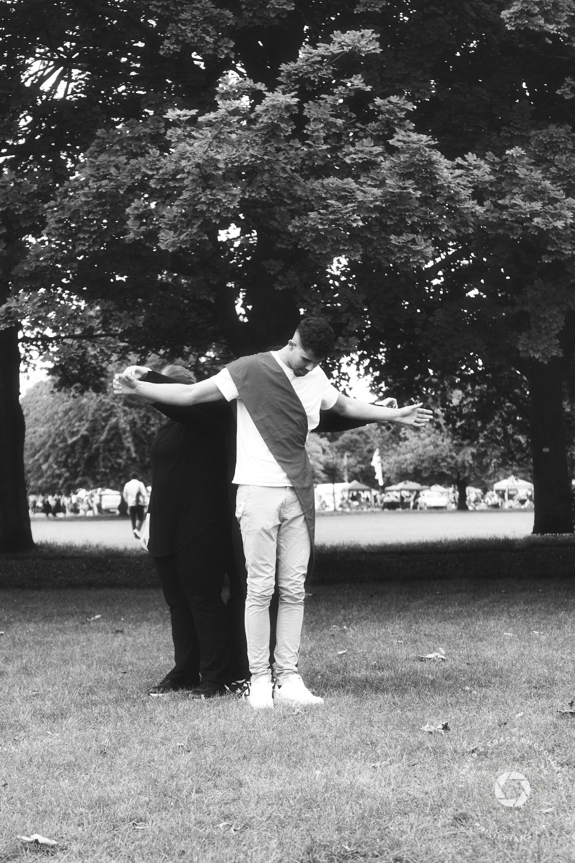 A black and white photo of two people standing on grass with trees in the background. One person is draped in a cloth and stands with outstretched arms, while the other stands close behind. The scene suggests an outdoor gathering or event.