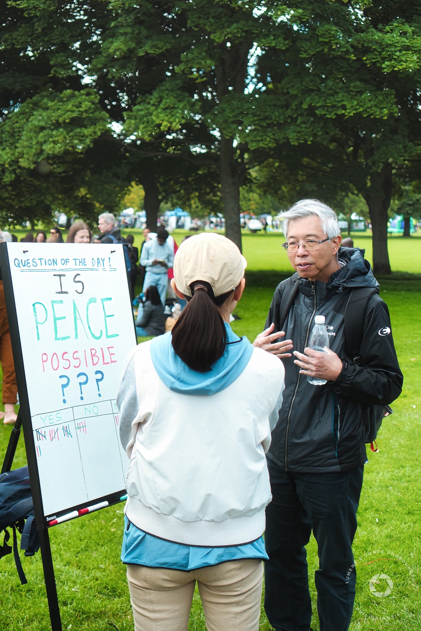 A person in casual clothing stands next to a whiteboard sign posing the question, "Is peace possible?" with tally marks under yes and no columns. Another person converses with them while holding a water bottle. The scene takes place in a park with pe