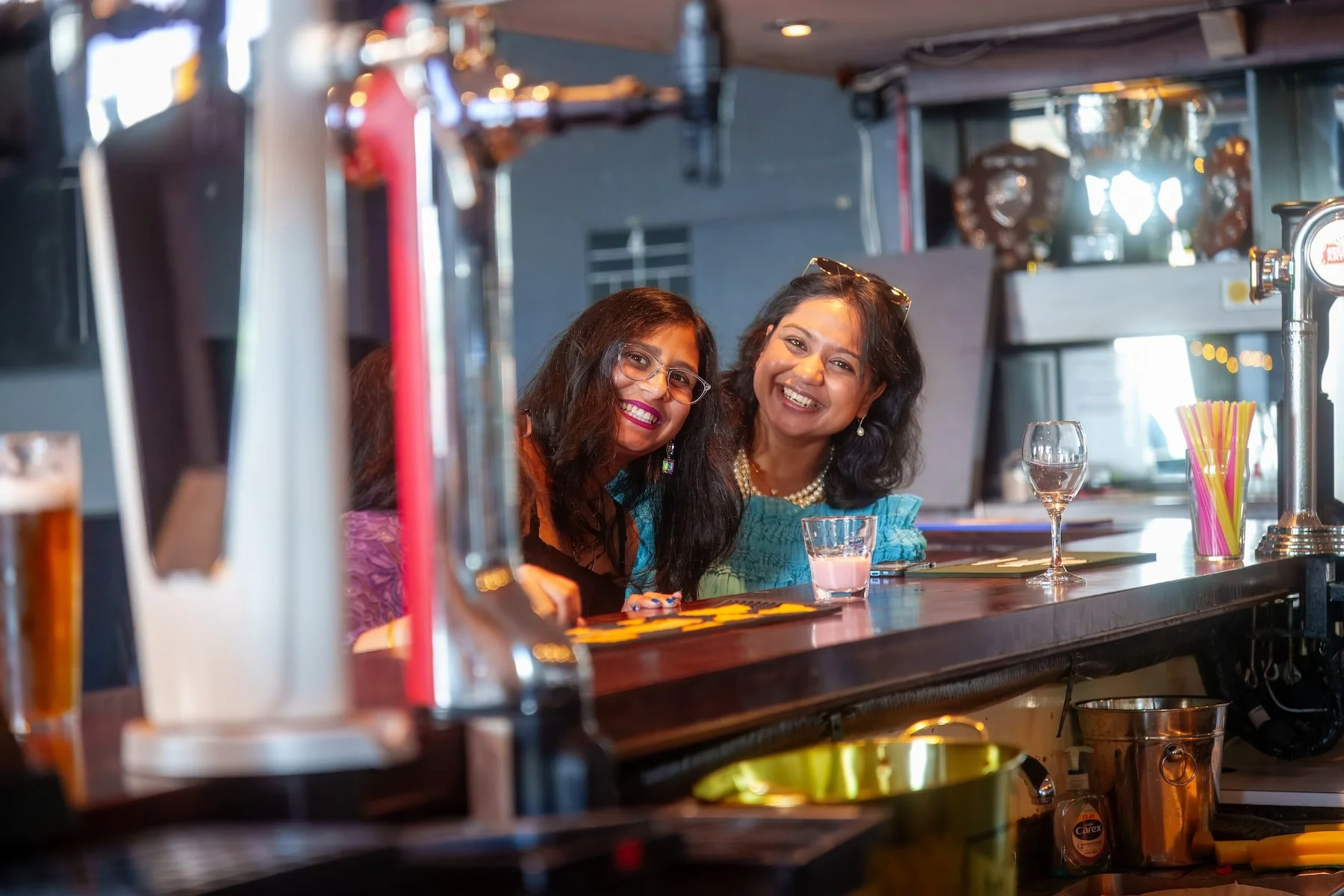 Two women smiling at a bar counter with drinks and colorful straws, behind the bar with shelves and trophies in the background.