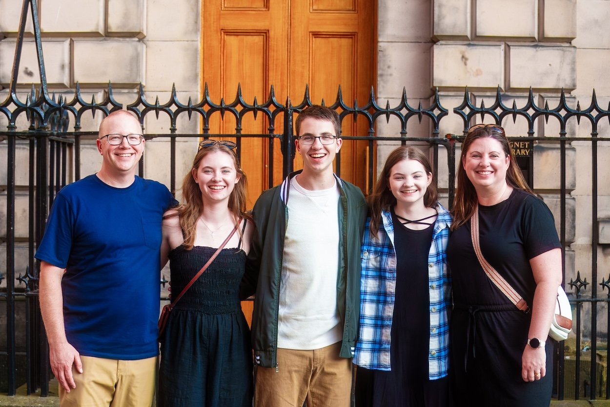 A group of five smiling people standing in front of a building with a black iron fence and a wooden door.