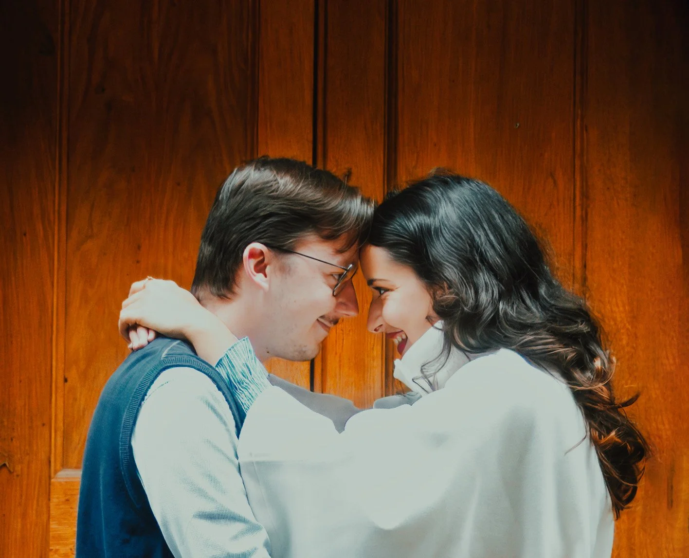 A young couple faces each other touching foreheads and smiling with joy, indoors against a wooden wall background.