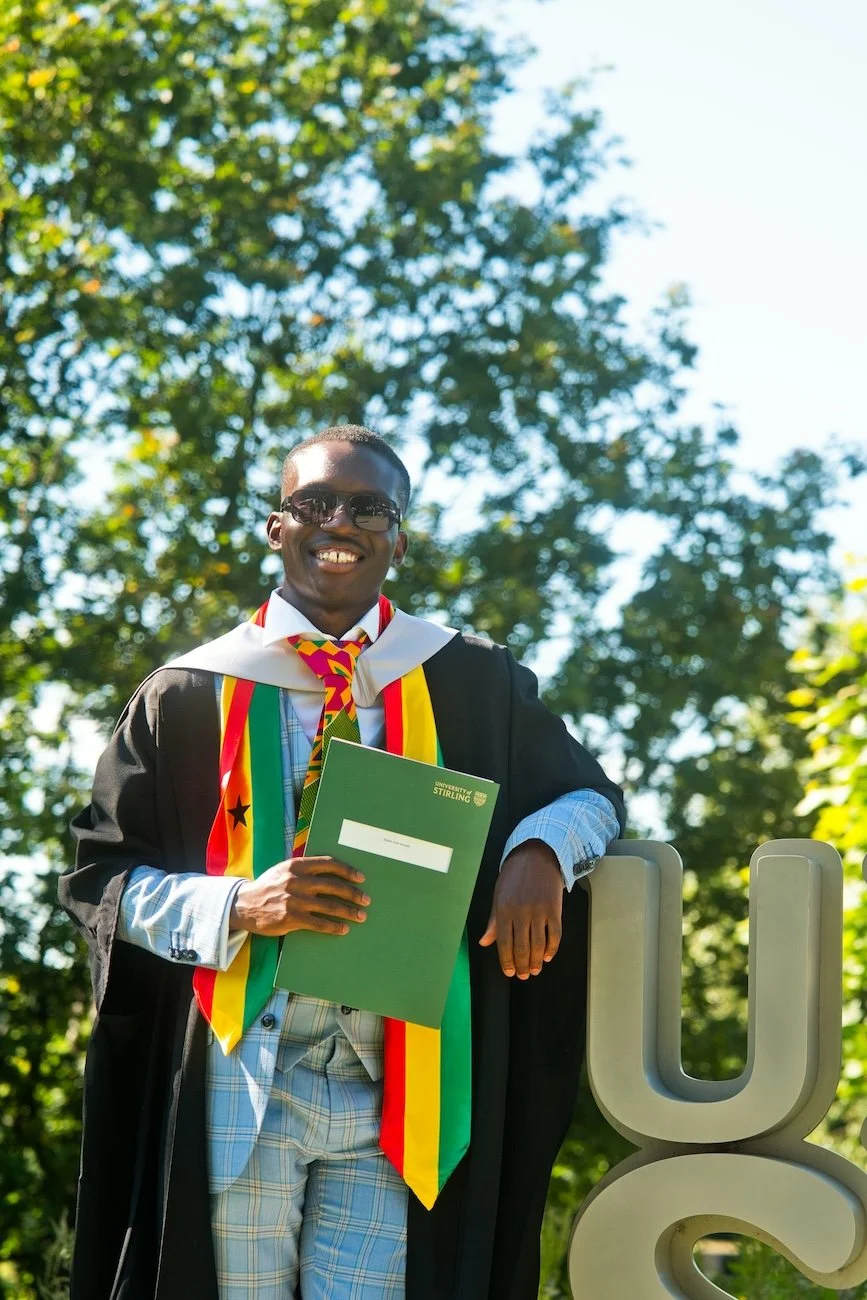 Graduation student in cap and gown holding diploma outdoors with trees in the background.