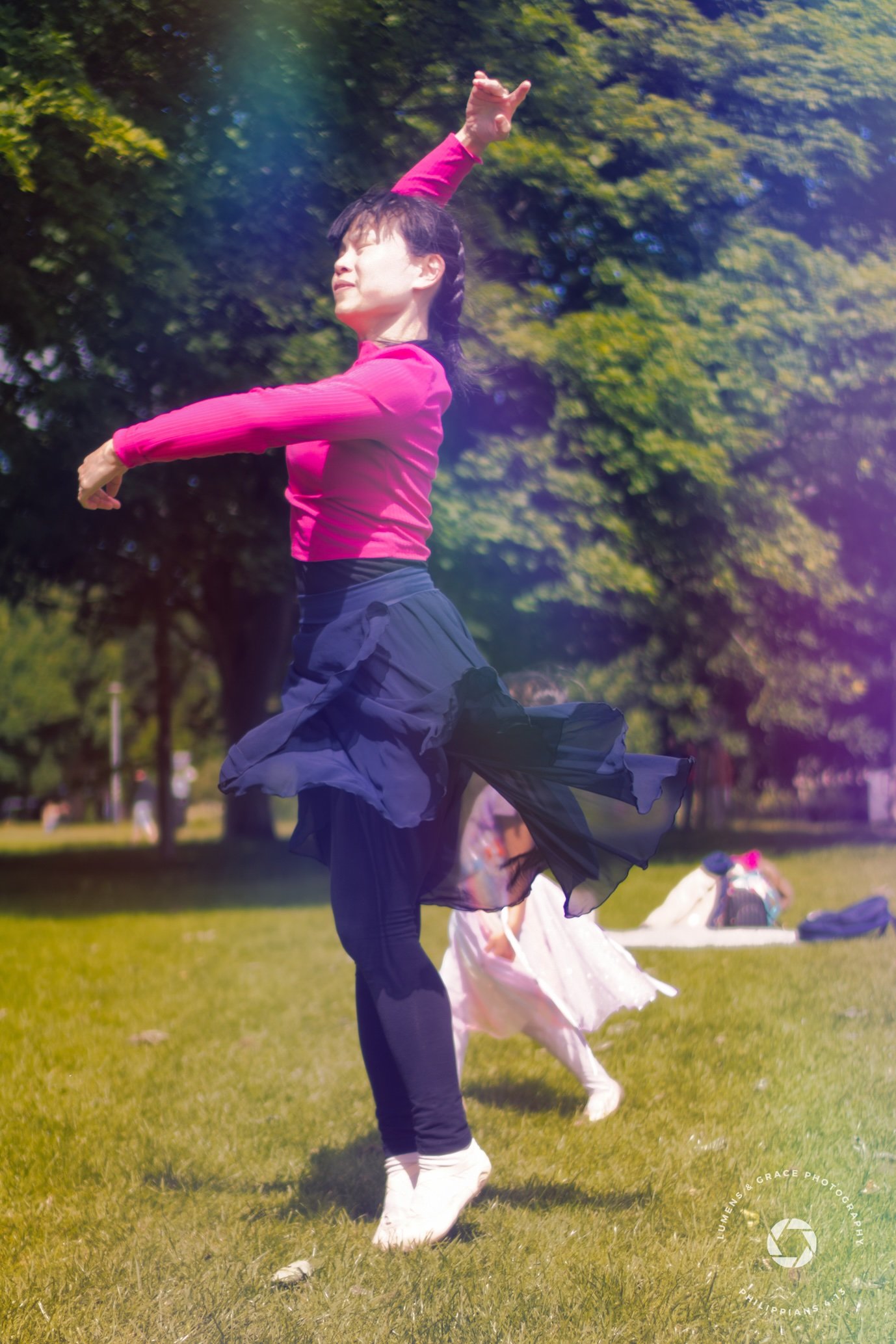 A person in a pink shirt and flowing skirt gracefully dancing outdoors on grass, with trees in the background.
