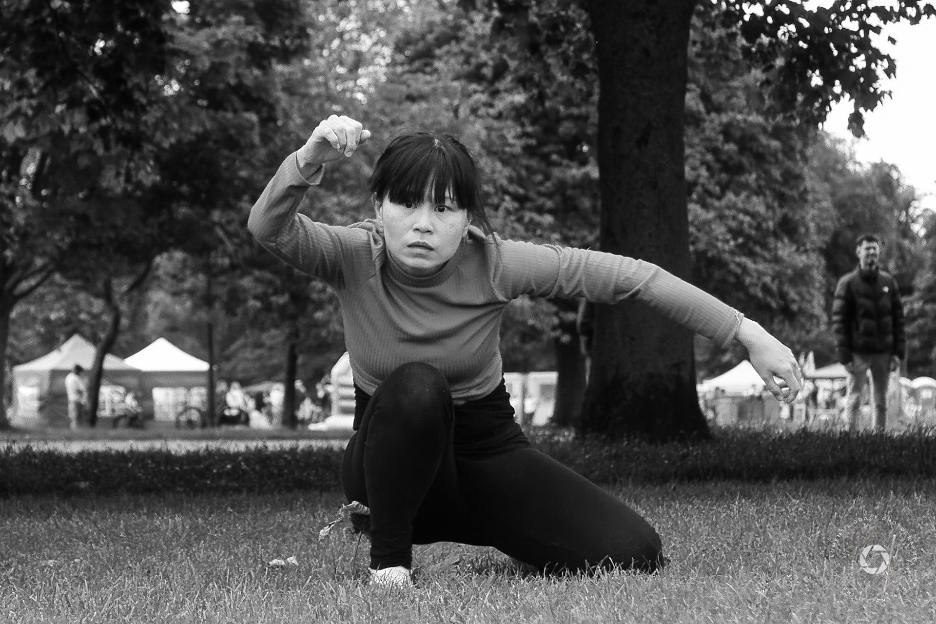 Woman in a kneeling pose with an arm extended outdoors, black and white photo, trees in the background.