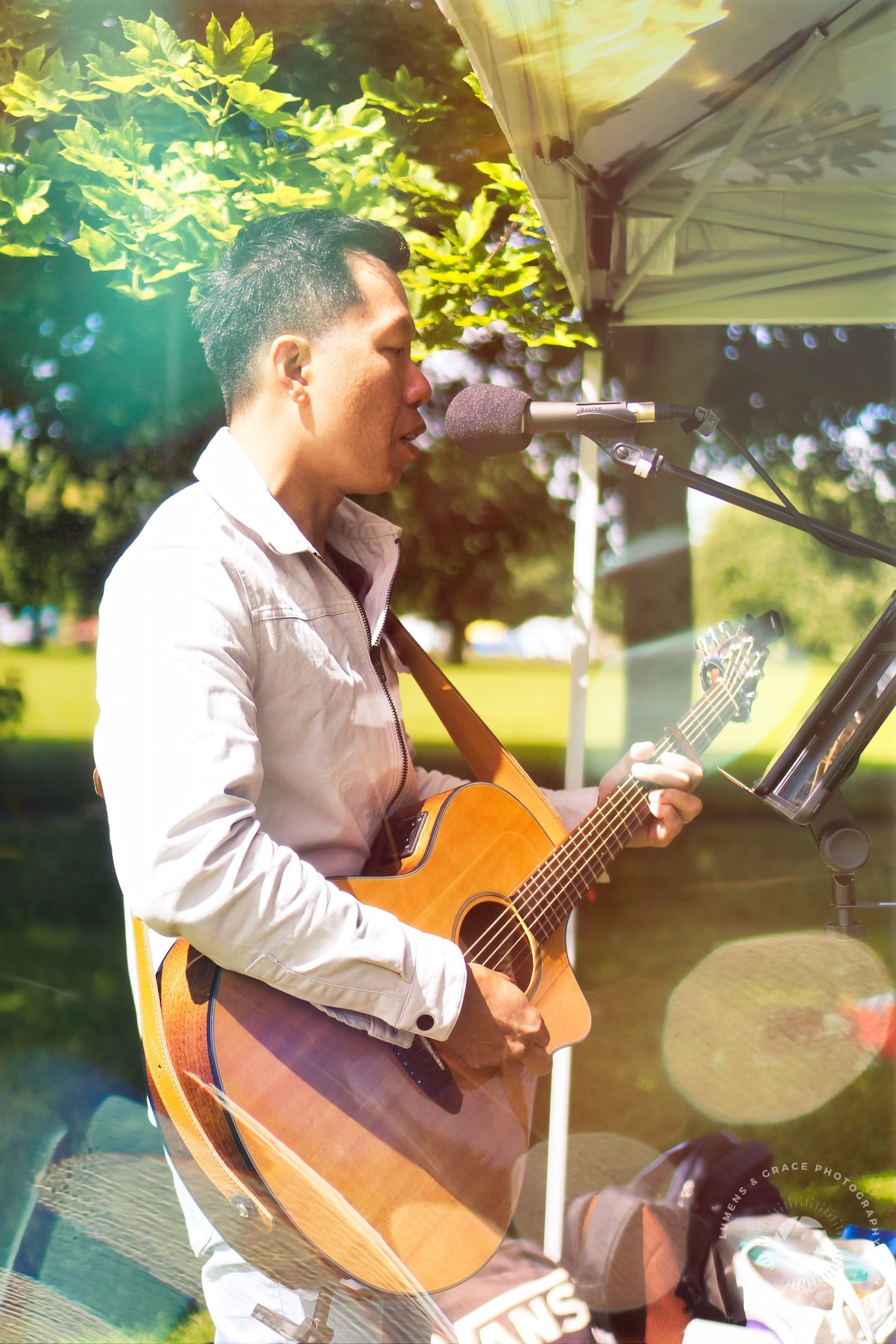 Musician playing an acoustic guitar and singing into a microphone outdoors under a canopy.